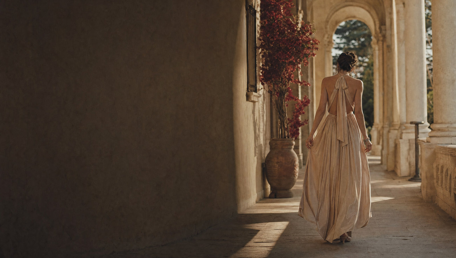 Woman in a flowing champagne occasion dress walking through a sunlit stone colonnade